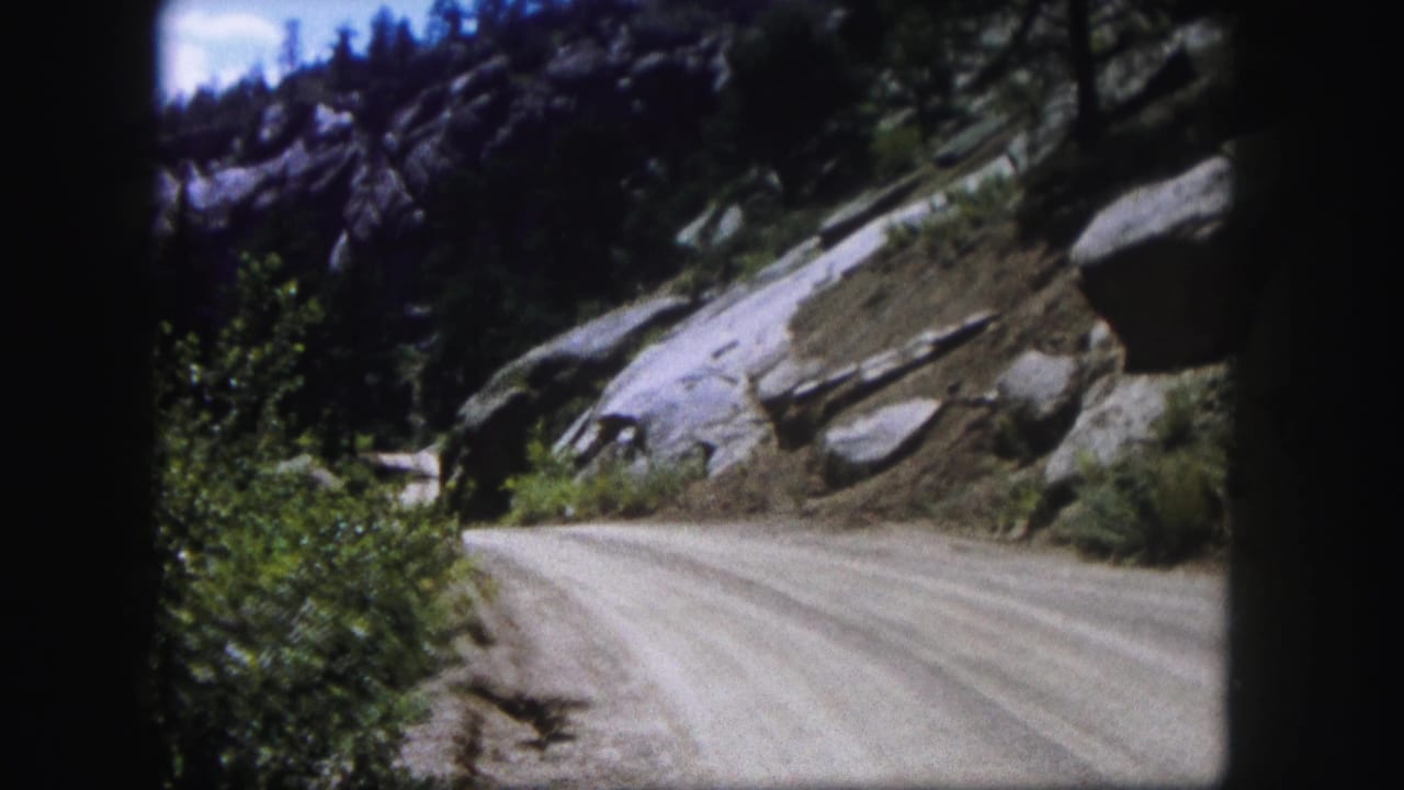 Curving dirt road with boulders, shrubs, rocky hillside, and trees.