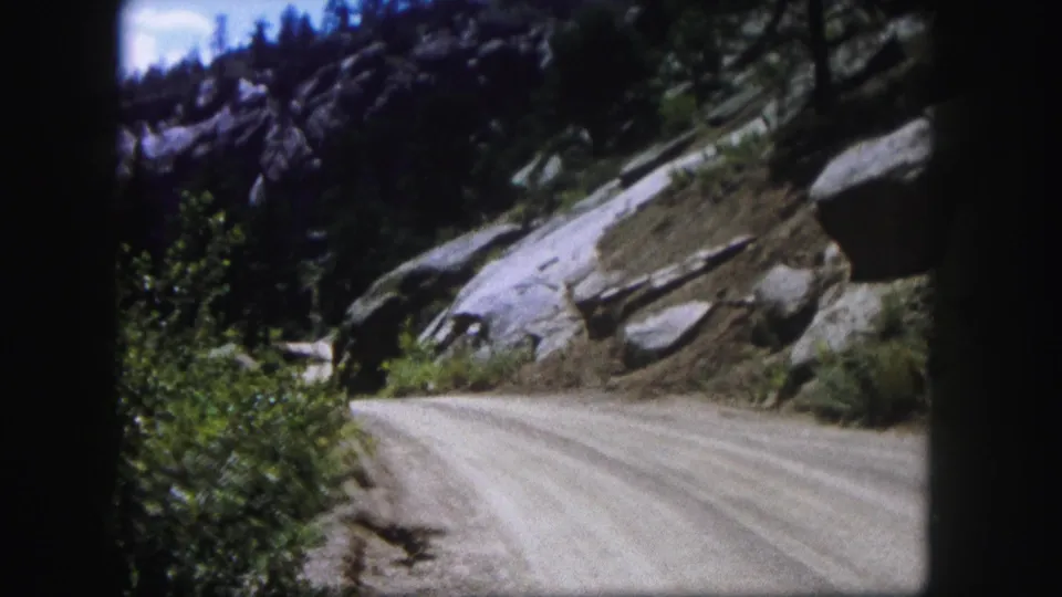 1958 Colorado home-movie frame with the road curving along a rocky slope lined by shrubs, boulders, and evergreens.