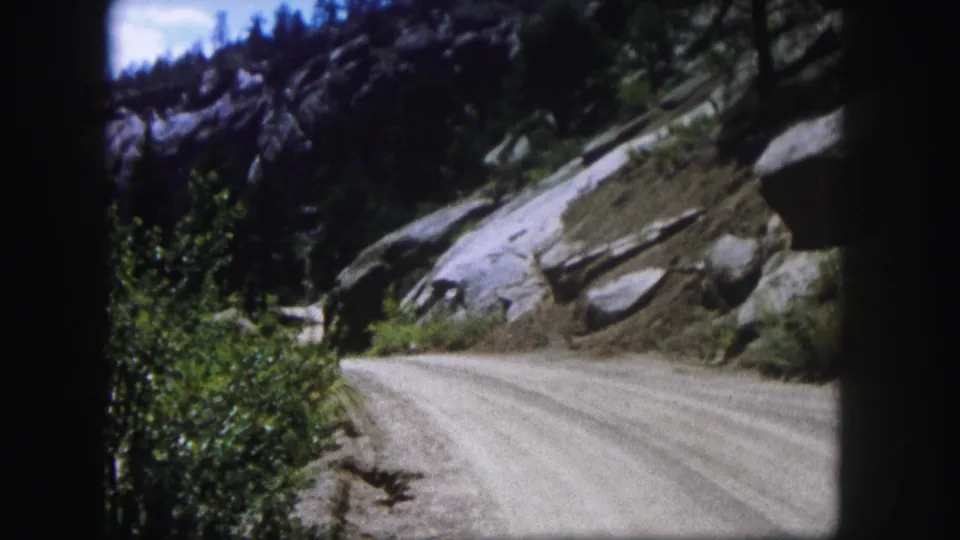 1958 Colorado 8mm frame looking along a dusty road bordered by brush, exposed rock, and dark evergreen trees.