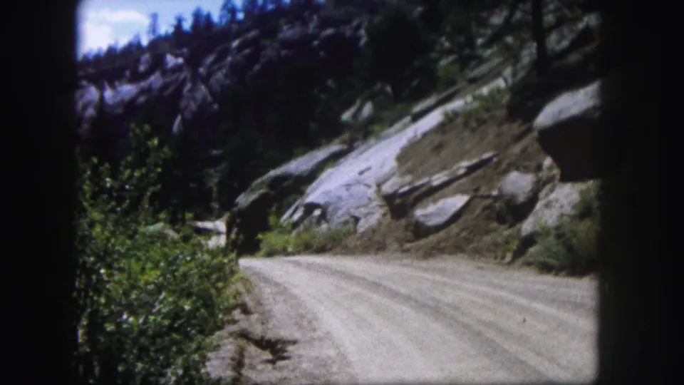 1958 Colorado frame showing a bright dirt road running past large boulders, scrub vegetation, and evergreen-covered rock.