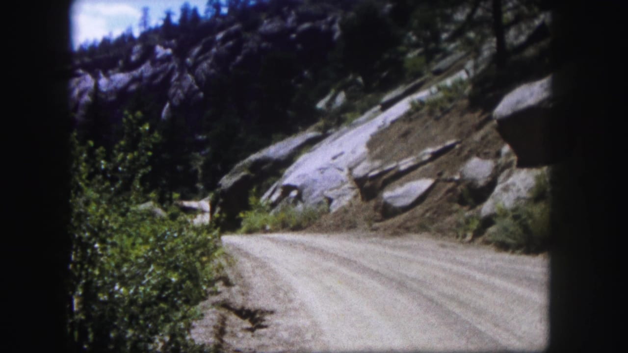 Colorado mountain road scene from 1958 8mm footage, with rocky slopes, evergreens, brush, and blue sky.