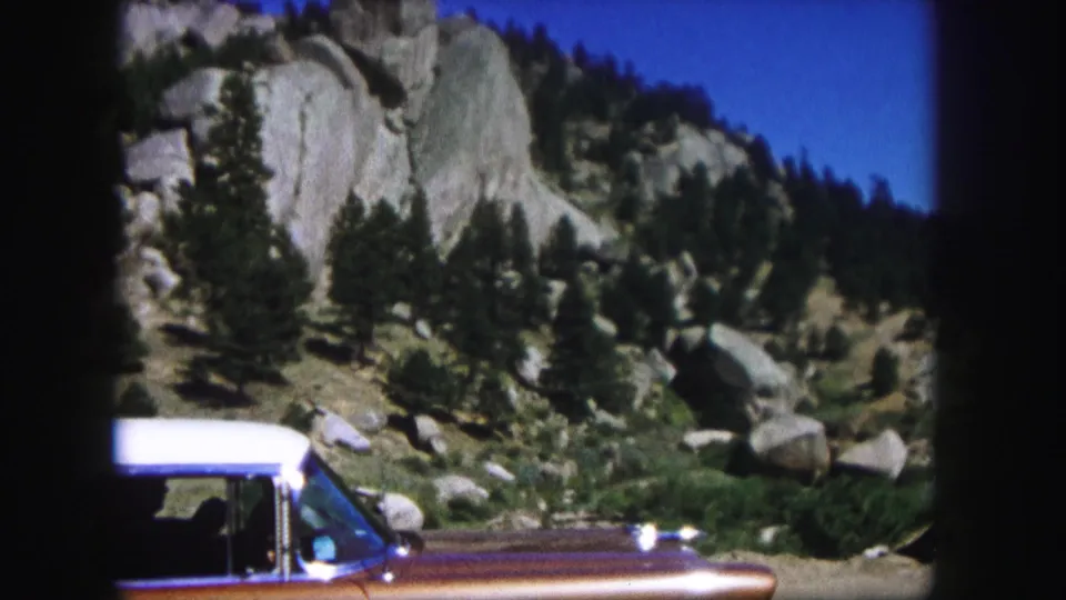 A partially framed brown-and-white vintage car appears at the edge of a 1958 Colorado mountain roadside view.
