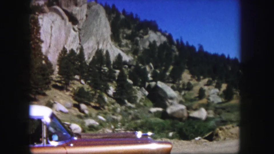 Close roadside view of a brown-and-white vintage car with Colorado rock formations and evergreens in the background.