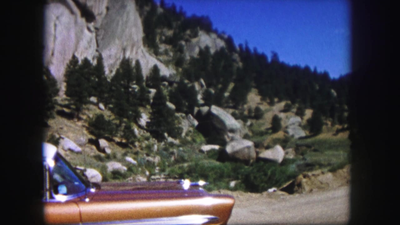 1958 Colorado 8mm frame showing the front half of a copper-and-purple vintage car parked along a dirt road below rocky slopes and evergreens.