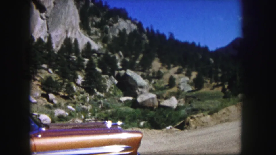 1958 Colorado home-movie frame with a vintage car partly cropped in the foreground and a rugged mountain slope, trees, boulders, and dirt road behind it.