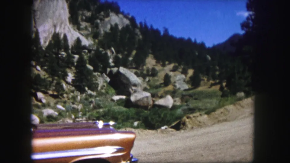1958 Colorado 8mm view of a copper vintage car cropped along the lower frame, with a dirt road, boulders, evergreens, and mountain slopes in the background.