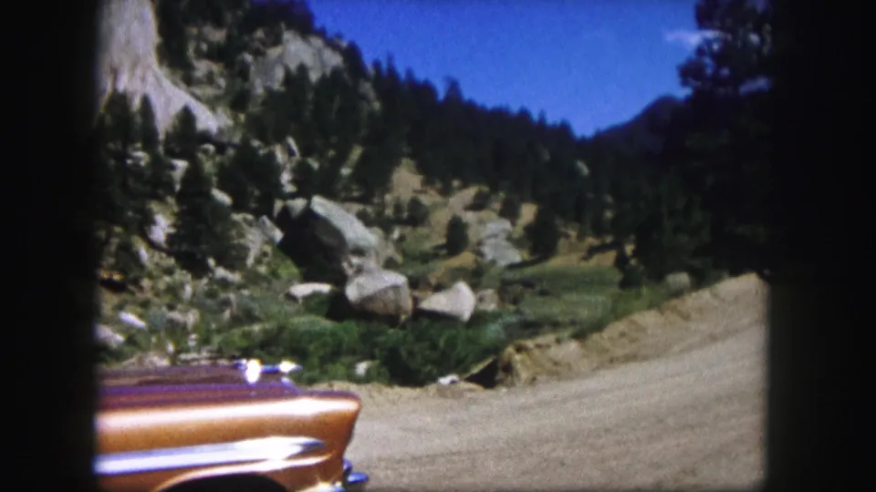 1958 Colorado frame showing the vintage car low in the foreground, with a dirt road curving past boulders, evergreens, and shadowed mountain slopes.