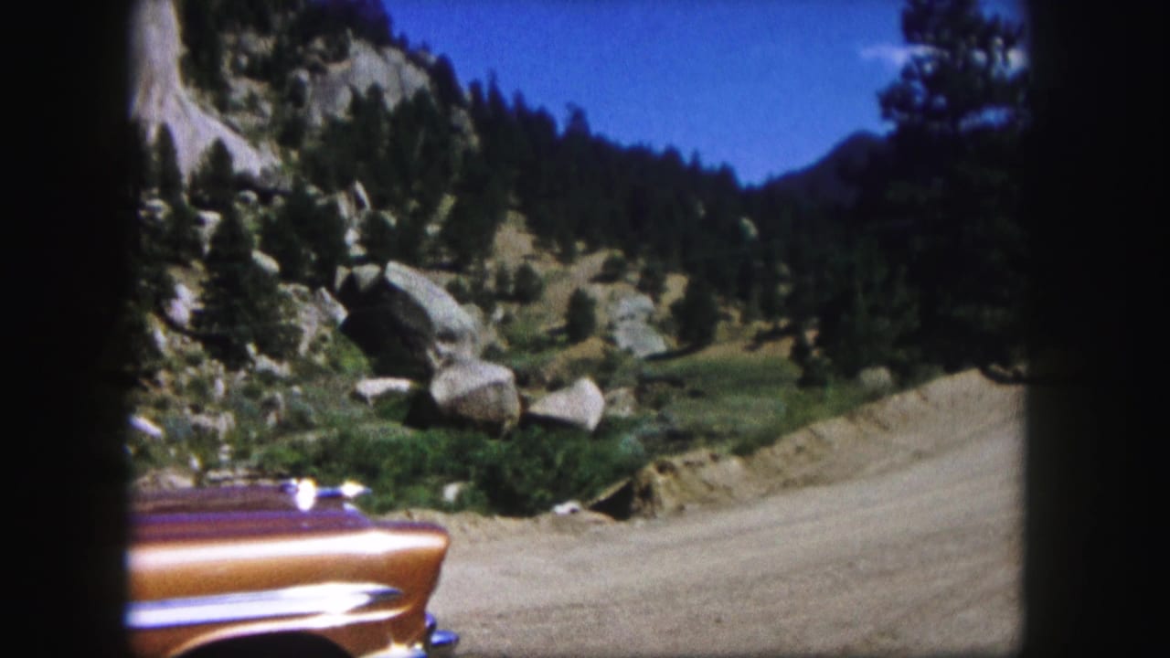 1958 Colorado 8mm frame showing a dirt mountain road, rocky slope, pines, blue sky, and part of an orange car fender.