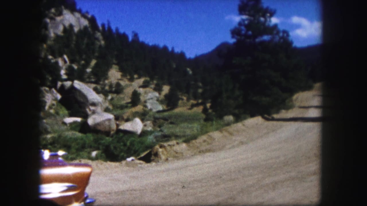 Mountain dirt road curving past rocks, pines, and the partial front of an orange car.
