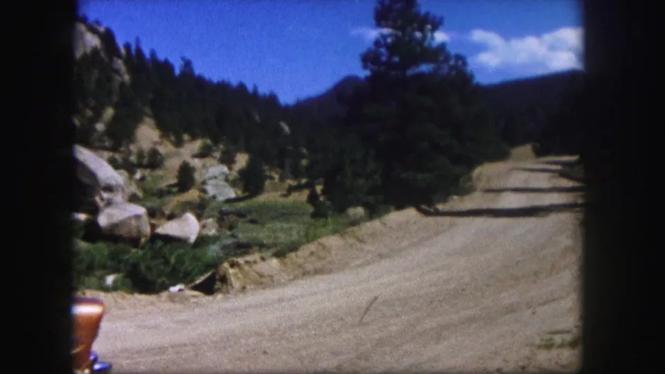 Colorado mountain road scene from 1958 8mm footage, showing a broad dirt road, pines, boulders, and a cropped orange car edge.