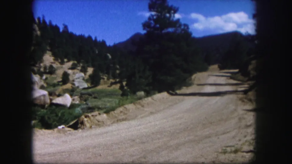 1958 Colorado 8mm frame of an empty dirt road continuing through pine trees, boulders, and mountain terrain.