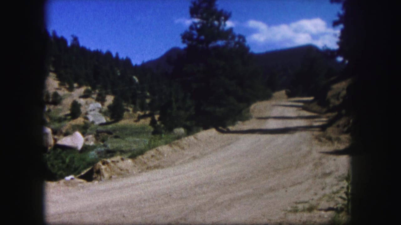 1958 Colorado 8mm frame showing an unpaved road curving through dry pine-covered mountain terrain under a bright sky.