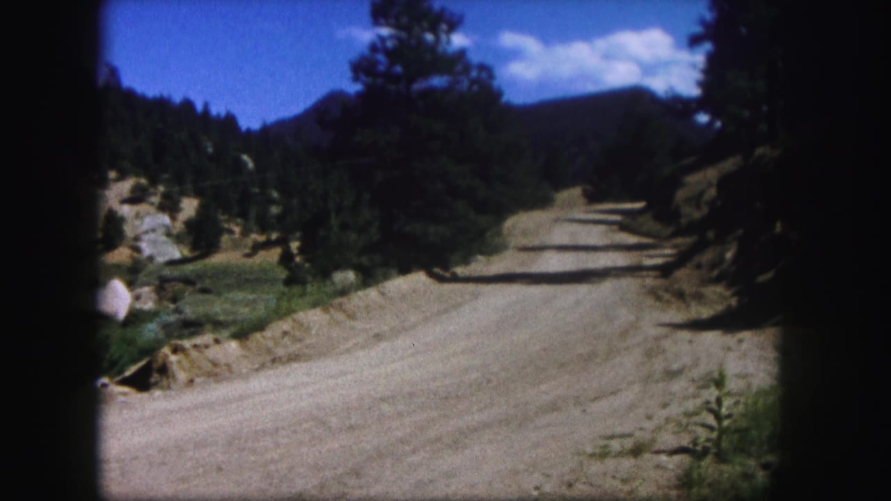 Unpaved road rises through pine forest and rocky mountain terrain beneath clouds.