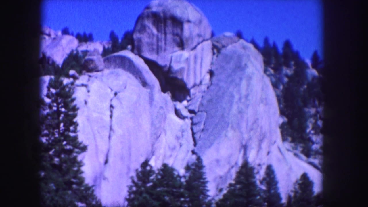 1958 Colorado 8mm frame showing pale granite formations, dark evergreen trees, and a saturated blue mountain sky.