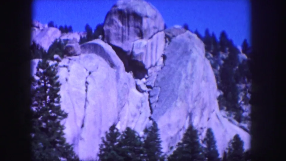 1958 Colorado 8mm frame showing pale granite formations, dark evergreen trees, and a saturated blue mountain sky.