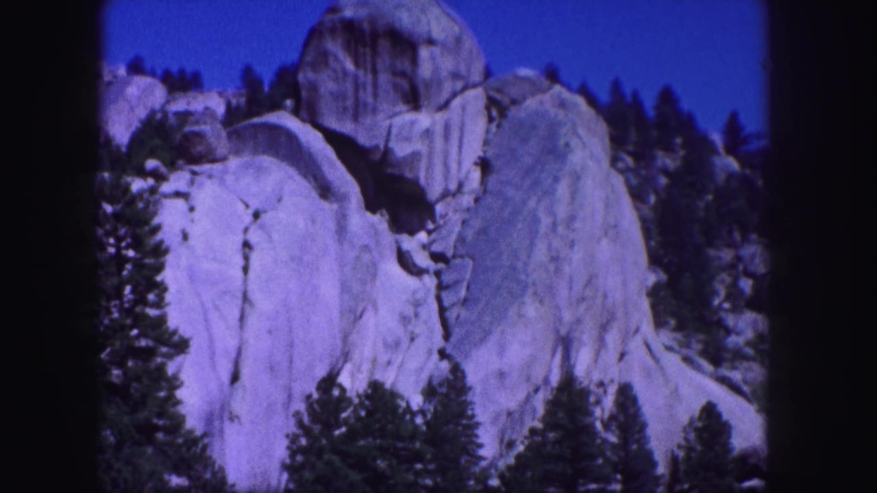 1958 Colorado home-movie frame with massive granite formations, tree silhouettes, and blue sky in soft 8mm color.