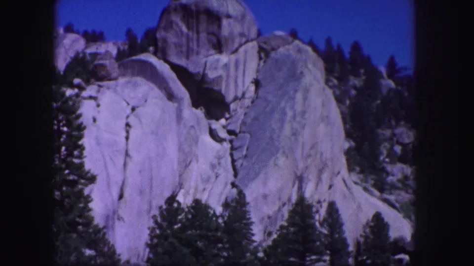 1958 Colorado mountain landscape frame showing granite rock faces, conifers, and a deep blue sky on 8mm film.