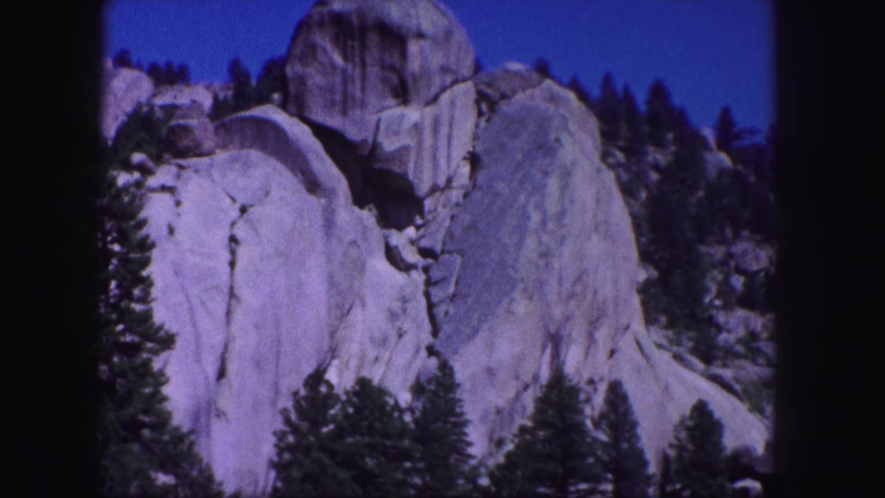 1958 Colorado 8mm frame showing pale granite rock formations rising behind evergreen trees beneath a saturated blue sky.