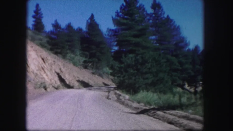 1958 Colorado travel frame showing a dirt road along a sloped cut bank with evergreen trees and roadside brush.
