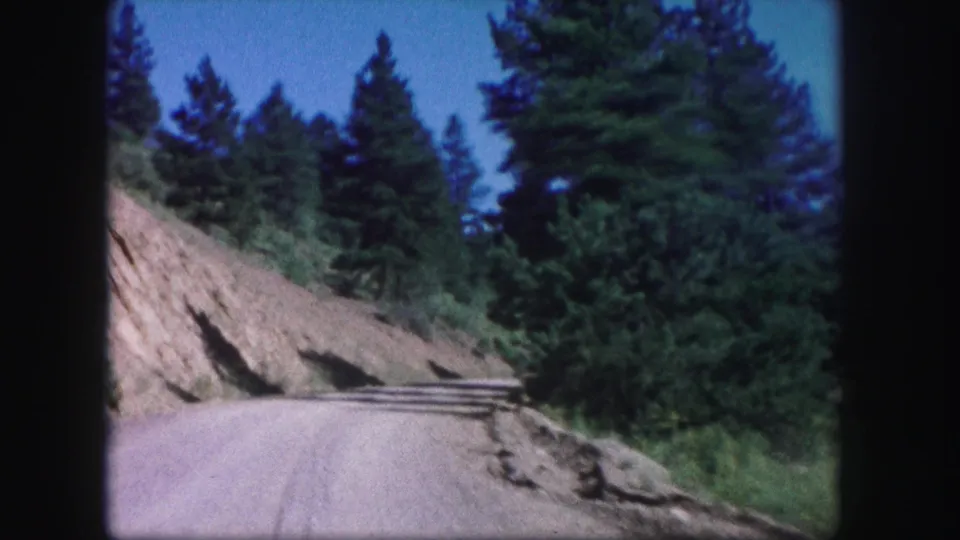 Archival 8mm Colorado road view with a pale unpaved road, hillside cut, dense conifers, and saturated mid-century color.