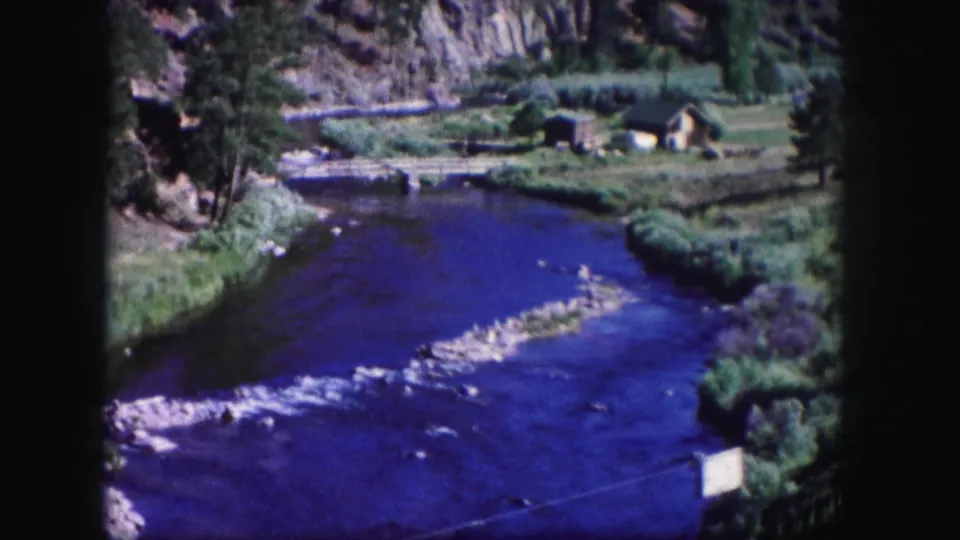 1958 Colorado 8mm frame showing a vivid blue river, rocky shallows, green banks, and a small riverside cabin in the distance.