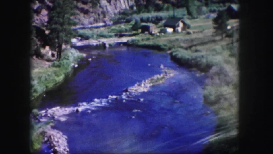 1958 Colorado 8mm frame looking over a blue river bend with rocky shallows, grassy banks, pine trees, and a small cabin beyond.