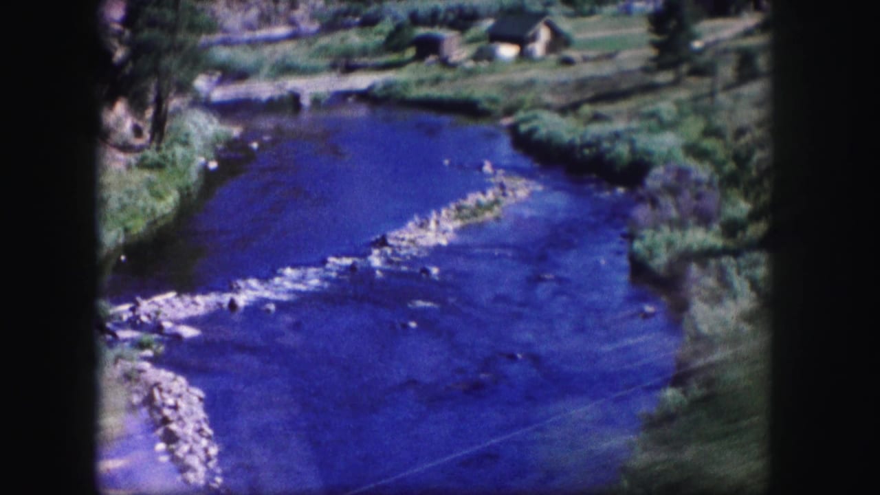 Blue river winding through green banks with a rocky riffle and small buildings in the distance.