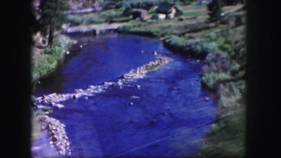 1958 Colorado 8mm frame showing a blue river, rocky shallows, green banks, trees, and small riverside buildings in the background.