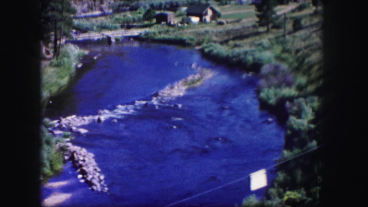 River bend with blue water, a pale gravel bar, green banks, trees, and small distant structures.