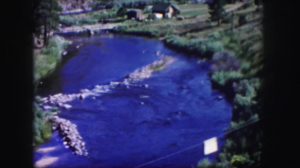 1958 Colorado home-movie frame of a river bend with blue water, gravel shallows, green banks, trees, and small buildings beyond.