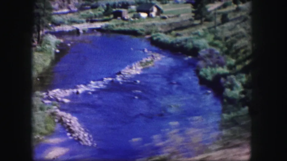 1958 Colorado 8mm frame showing saturated blue river water, rocky shallows, green vegetation, trees, and distant small buildings.