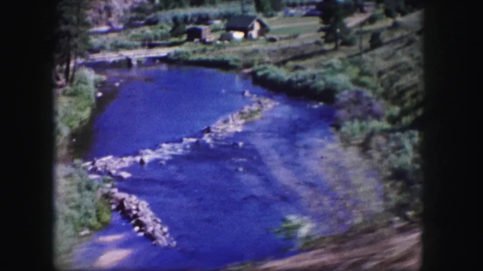 1958 Colorado home-movie frame with a wide blue river, pale gravel riffle, green banks, trees, and small cabin-like buildings in the distance.