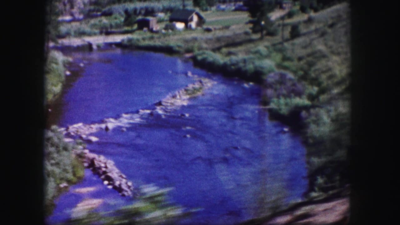 1958 Colorado 8mm frame showing a blue river curving through green banks with rocks in the water and a distant upper bank.