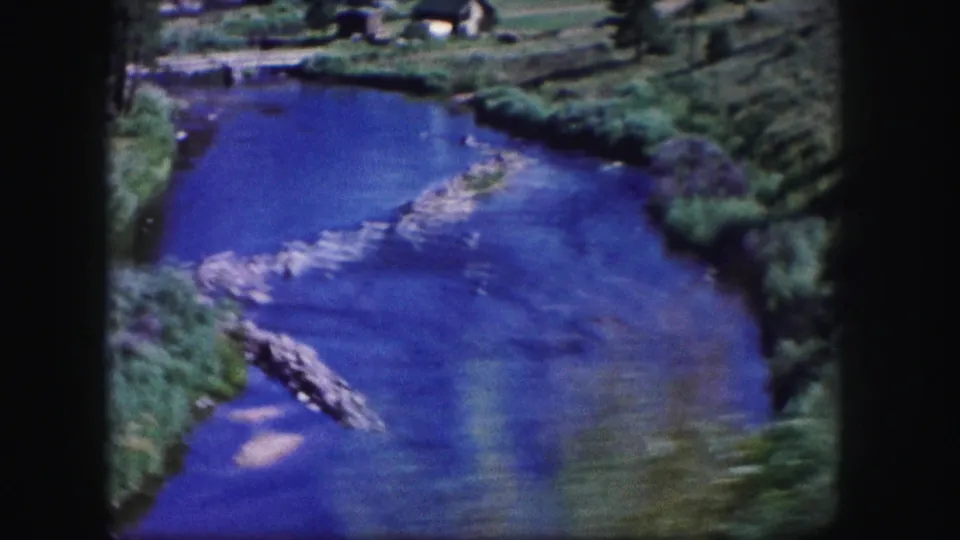 1958 Colorado home-movie frame of a blue river channel bordered by grass and low vegetation, with rocks breaking the current.