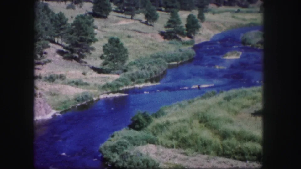 1958 Colorado 8mm view across a winding blue river, open grassy hills, scattered trees, and reed-like bank vegetation.