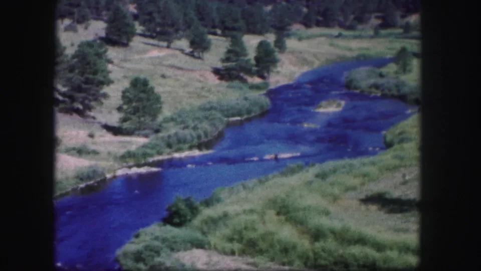 1958 Colorado home-movie frame showing a blue river below open grassy slopes with scattered trees and pale bank or path lines.
