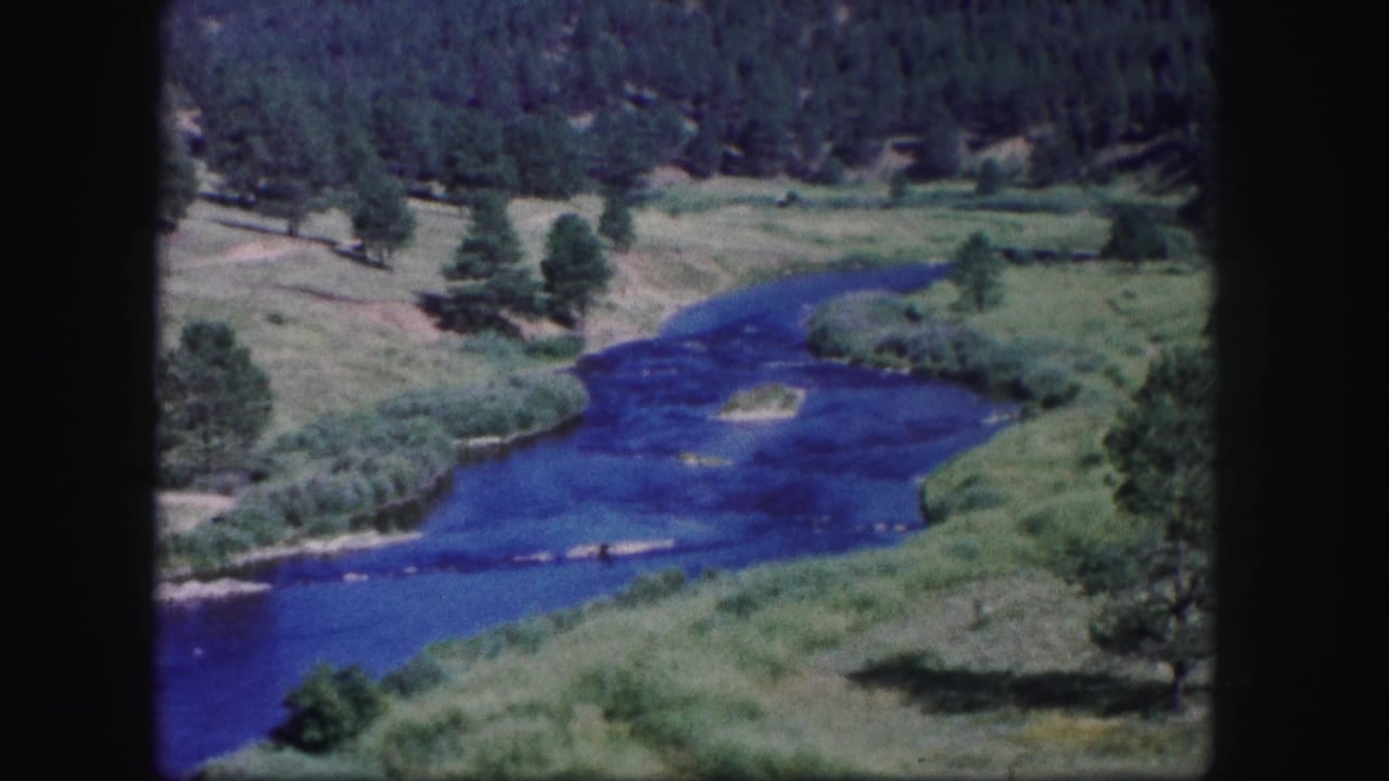 1958 Colorado landscape frame with a river bend, pale sandbar, grassy banks, scattered pines, and a wooded hillside.