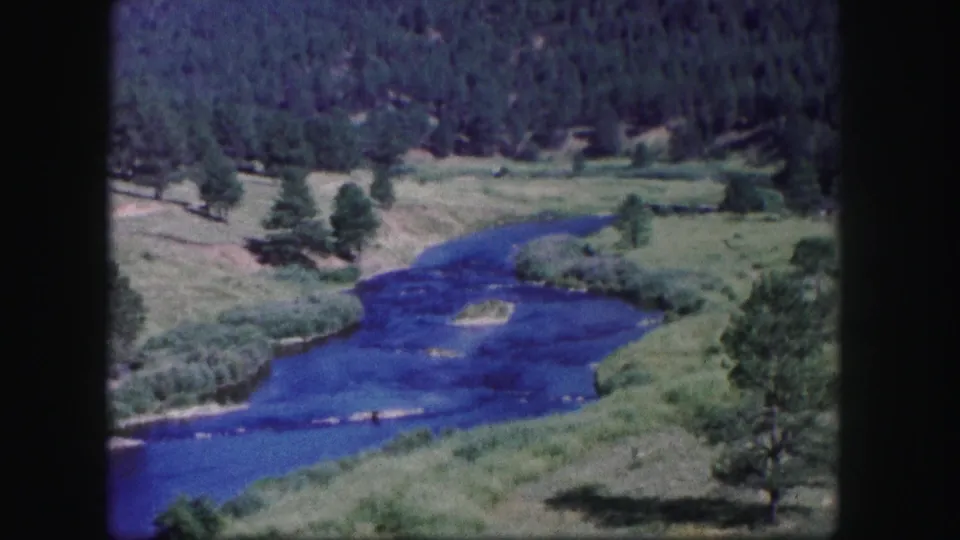 1958 Colorado 8mm view of a blue river winding across a green valley with scattered trees and dense forest behind.