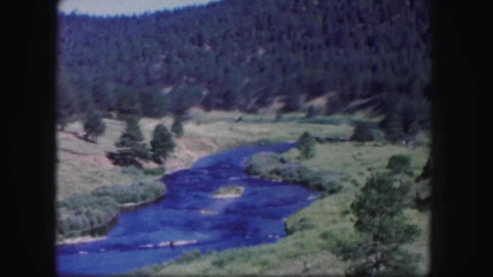 1958 Colorado frame with soft 8mm color, showing a blue river through meadowland with pines and dark forested hills.