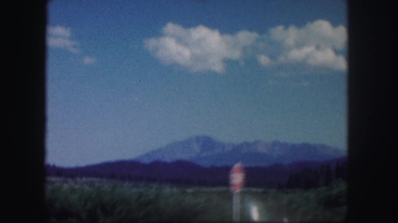 1958 Colorado travel view with distant mountains, blue sky, clouds, dark treeline, and a blurred roadside sign shape.