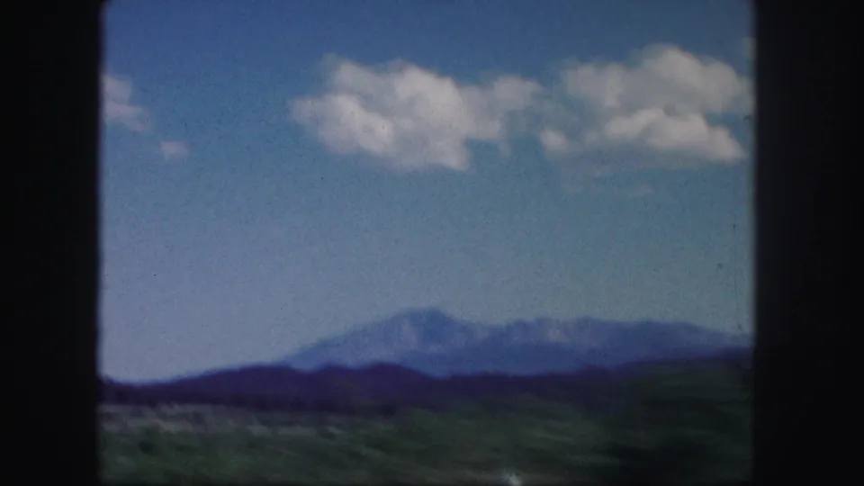 Soft 8mm Colorado landscape frame with a distant mountain range, low green foreground, and scattered clouds overhead.