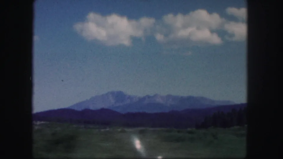 1958 Colorado 8mm frame showing distant mountains, a broad green foreground, dark treeline, and scattered clouds.