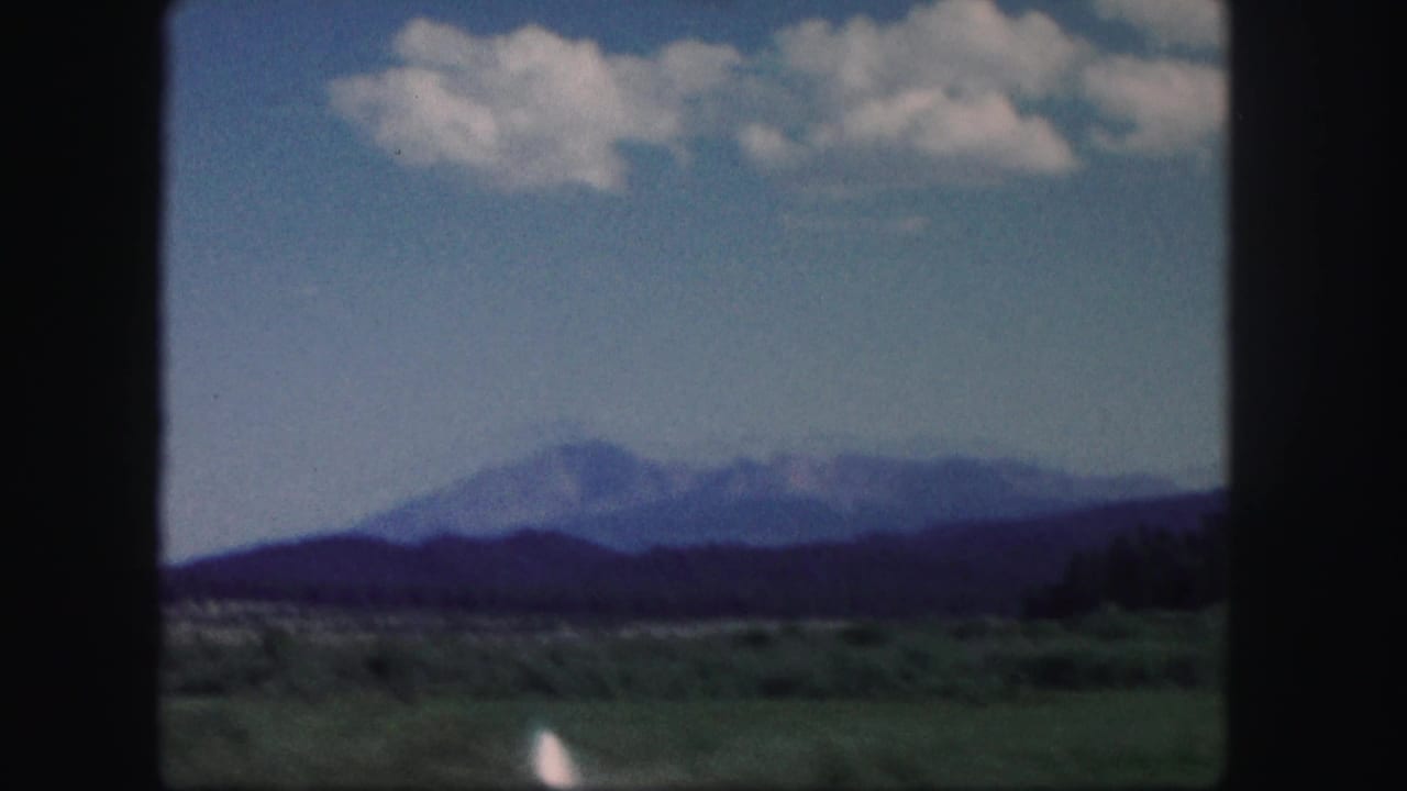 1958 Colorado 8mm frame showing distant mountains, green foreground fields, and scattered clouds from a moving travel viewpoint.