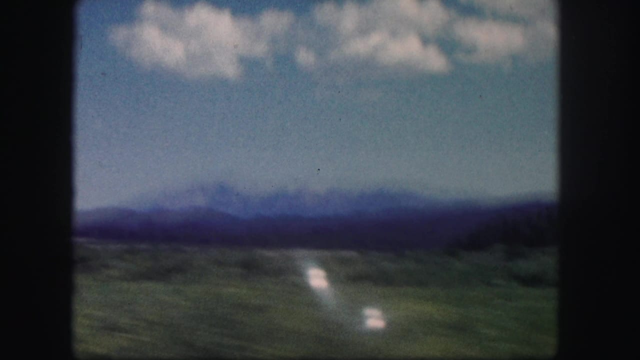 1958 Colorado home-movie frame with a blurred mountain horizon, green fields, blue sky, and bright reflective streaks from travel motion.