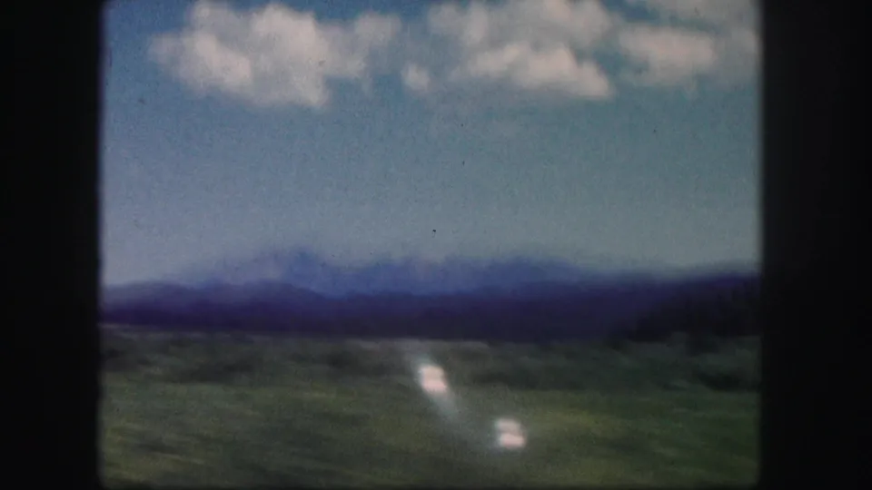 1958 Colorado home-movie frame with a blurred mountain horizon, green fields, blue sky, and bright reflective streaks from travel motion.