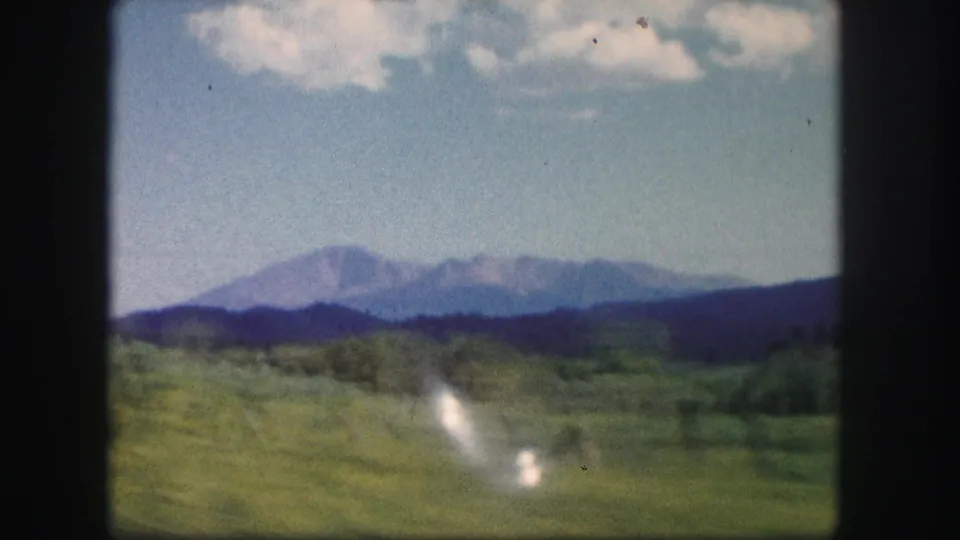 1958 Colorado travel frame with rolling green fields, a distant mountain range, scattered clouds, and bright reflective streaks.
