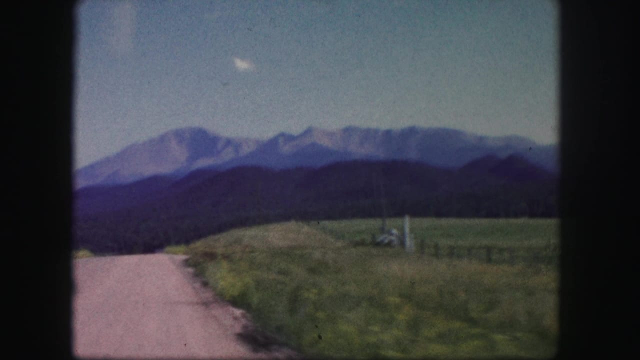1958 Colorado home-movie frame showing a pale rural road, grassy roadside, dark foothills, and a mountain range under blue sky.