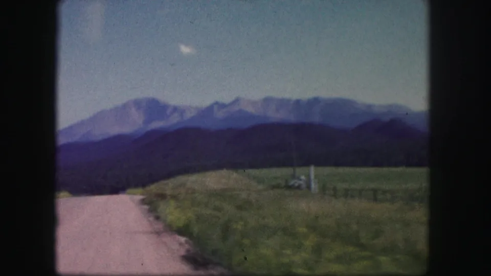 1958 Colorado home-movie frame showing a pale rural road, grassy roadside, dark foothills, and a mountain range under blue sky.