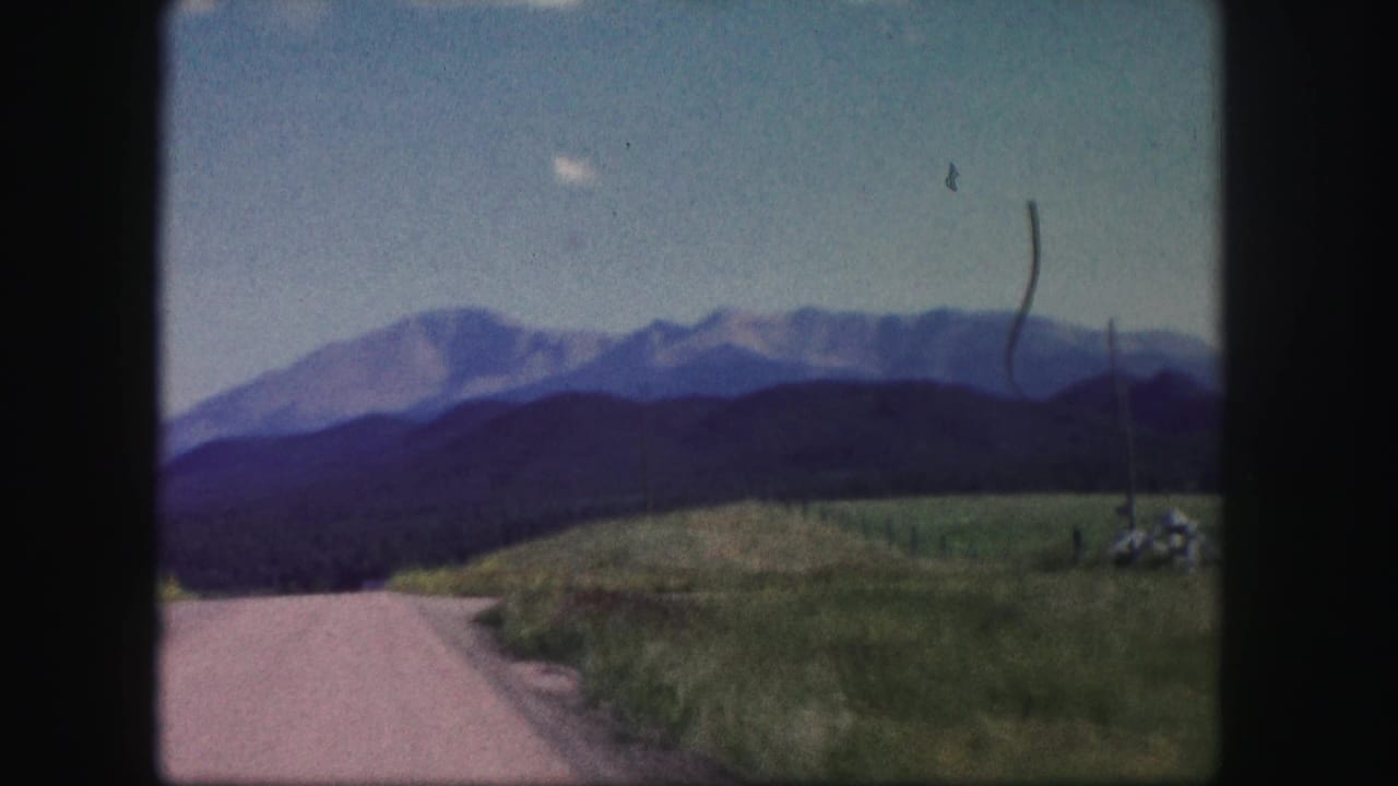 Curving roadside with grassy hillside, utility pole, forested ridge, and mountains.