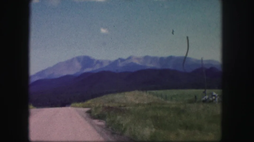 1958 Colorado road-trip view with a curving rural road, sloped grass, utility pole, dark tree line, and distant mountain range.